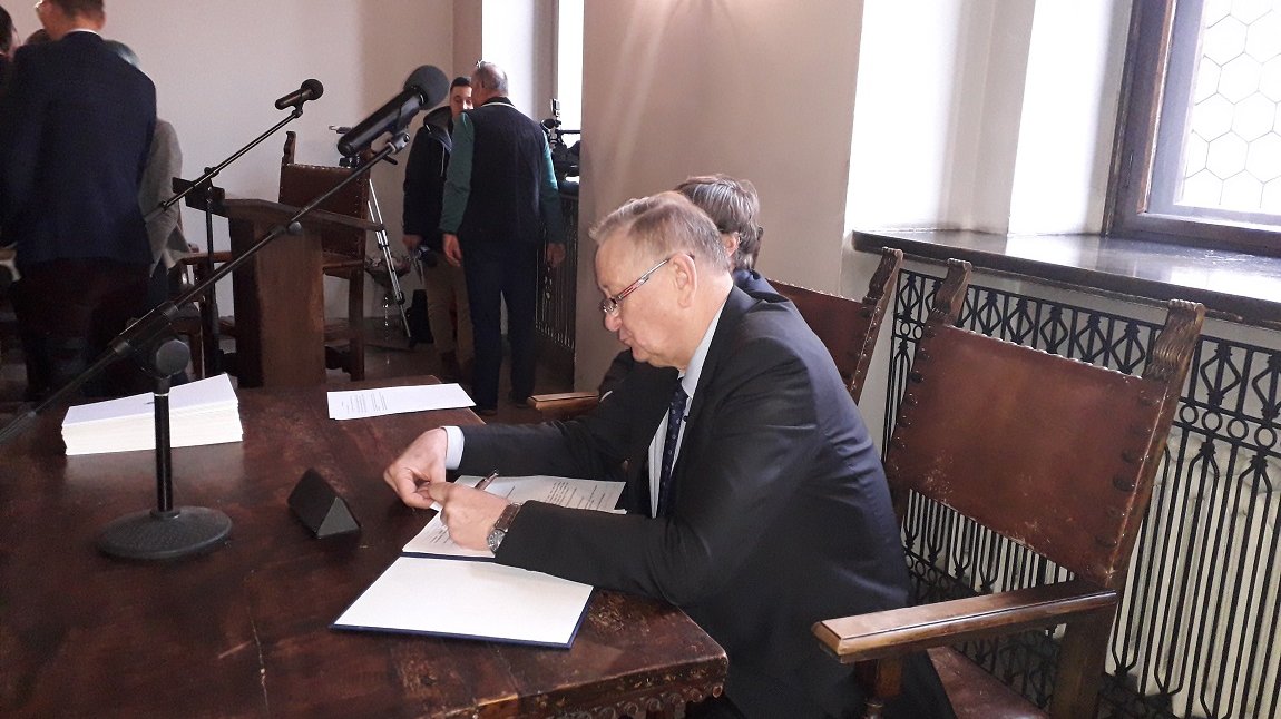 mężczyzna ubrany w garnitur siedzi przy stole i pisze coś na kartce, za nim mikrofony, dalej stoi kilka innych osób