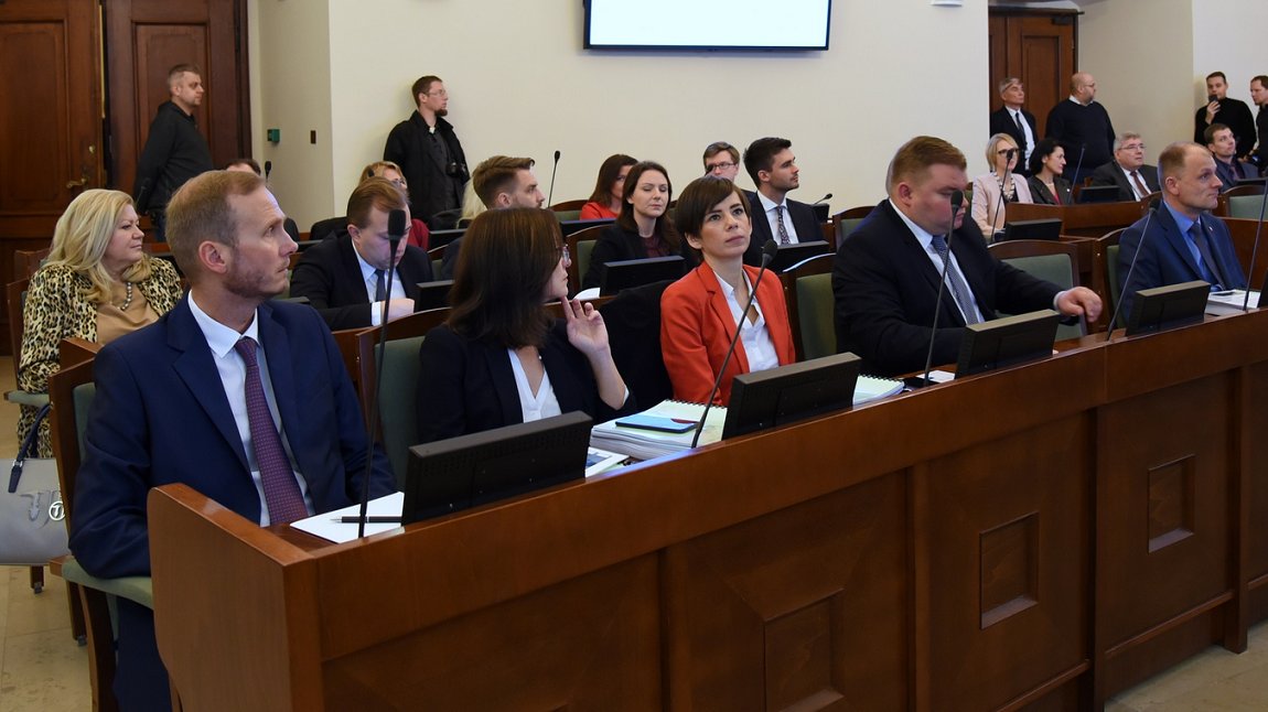 grupa osób kobiet i mężczyzn siedzi w rzędach w ławach, przy stanowiskach z mikrofonami i monitorami, za nimi pod ścianą kilka osób stoi