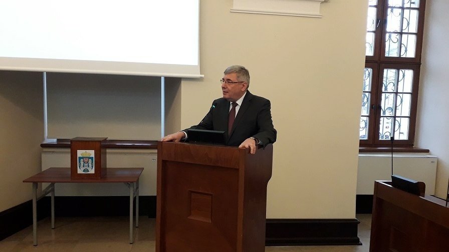 mężczyzna w garniturze i okularach stoi przy mównicy w sali o kremowych ścianach