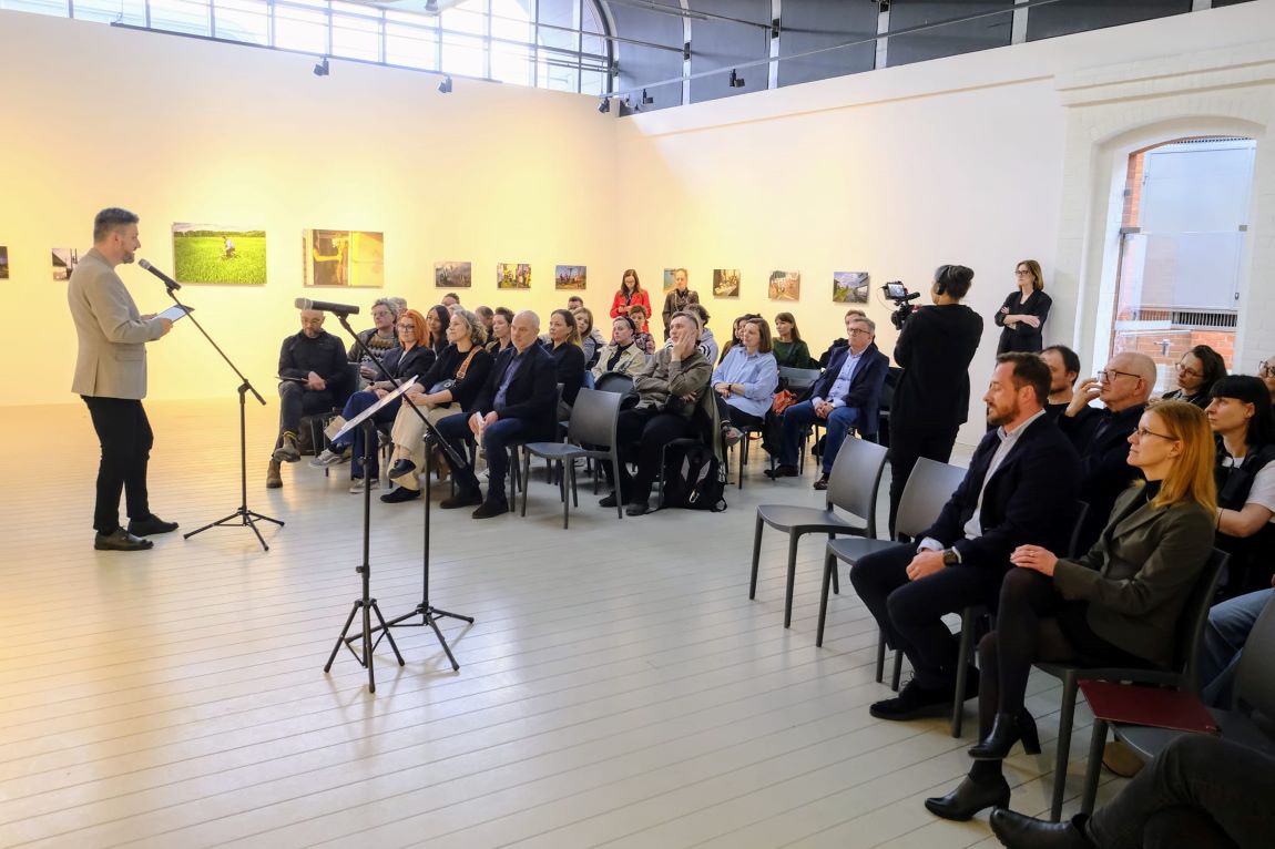 Sala cała w białym kolorze, na krzesłach ustawiony w rzędach siedzi kilkadziesiąt osób, kobiet i mężczyzn w różnym wieku, jeden mężczyzna w marynarce stoi na przeciwko nich i mówi do mikrofonu - grafika artykułu