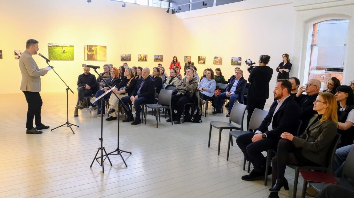 Sala cała w białym kolorze, na krzesłach ustawiony w rzędach siedzi kilkadziesiąt osób, kobiet i mężczyzn w różnym wieku, jeden mężczyzna w marynarce stoi na przeciwko nich i mówi do mikrofonu