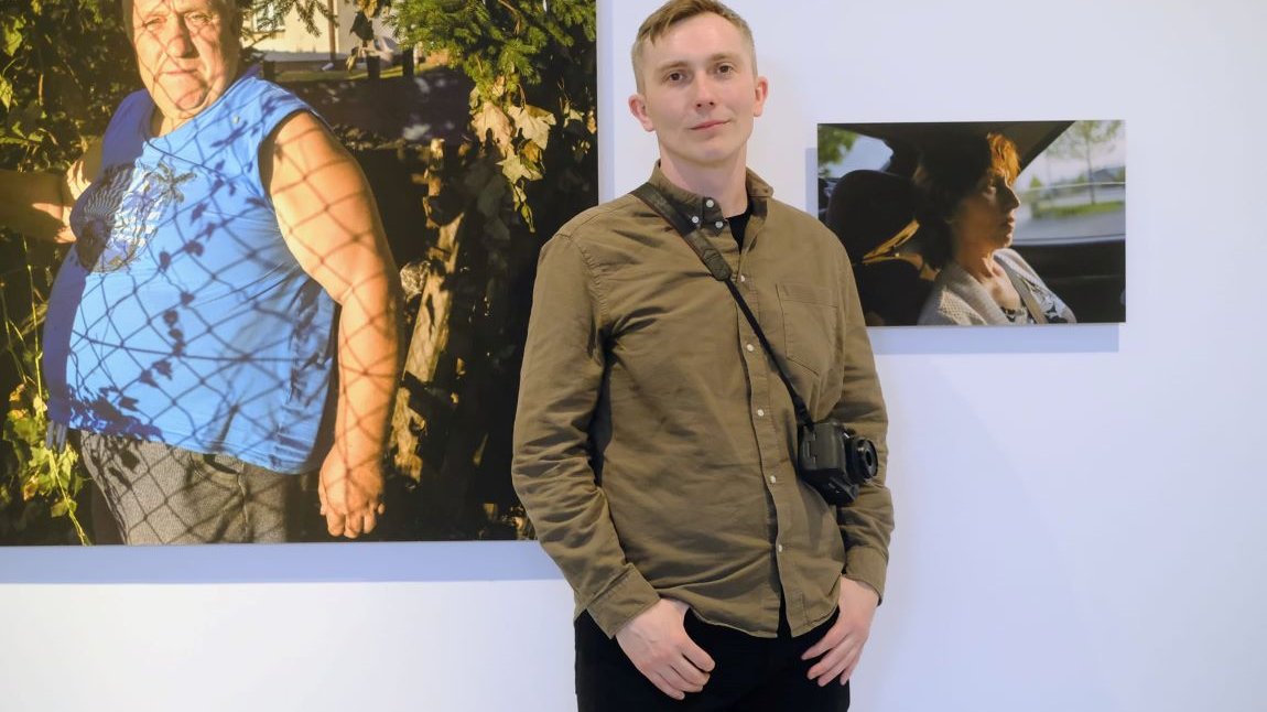 Młody mężczyzna o krótkich włosach, w koszuli, z aparatem fotograficznym przewieszonym przez ramię, za nim na ścianie wiszą dwie fotografie, na jednej z nich jest starszy mężczyzna, na drugiej kobieta w średnim wieku