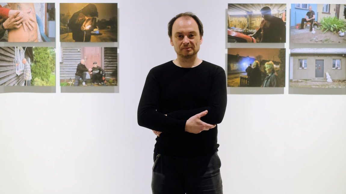 Mężczyzna w średnim wieku, ubrany cały na czarno, ciemne krótkie włosy, z przodu lekko łysiejący, za nim na ścianie wisi osiem fotografii przedstawiających sceny z życia mieszkańców baraków