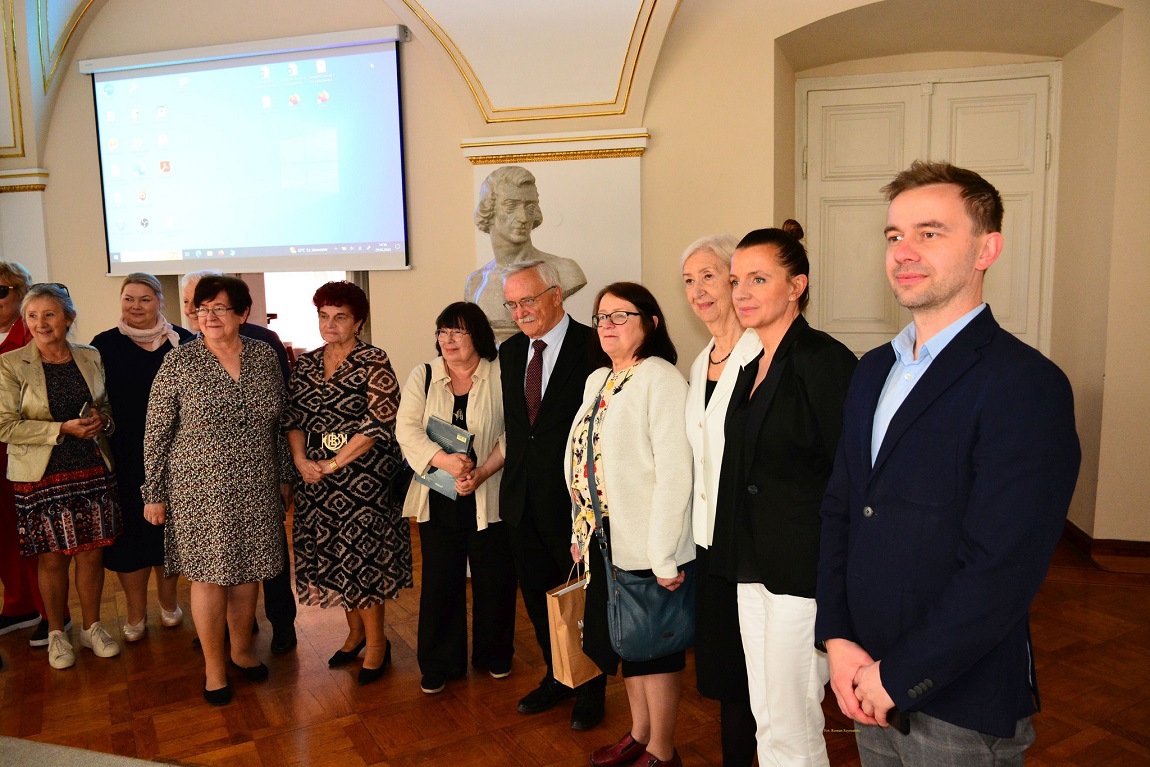 około kilkunastu osób w różnym wieku, kobiet i mężczyzn, stoi w szeregu, w sali o białych zdobionych ścianach - grafika artykułu