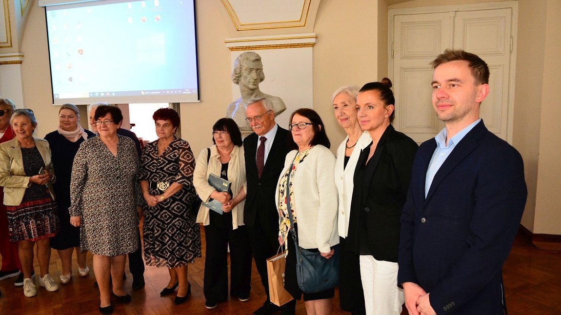 około kilkunastu osób w różnym wieku, kobiet i mężczyzn, stoi w szeregu, w sali o białych zdobionych ścianach