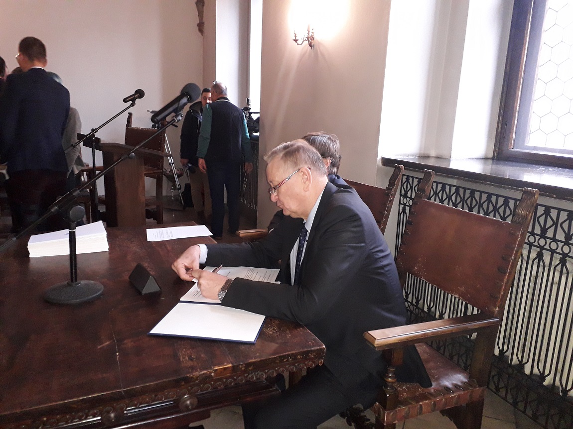 mężczyzna ubrany w garnitur siedzi przy stole i pisze coś na kartce, za nim mikrofony, dalej stoi kilka innych osób - grafika artykułu