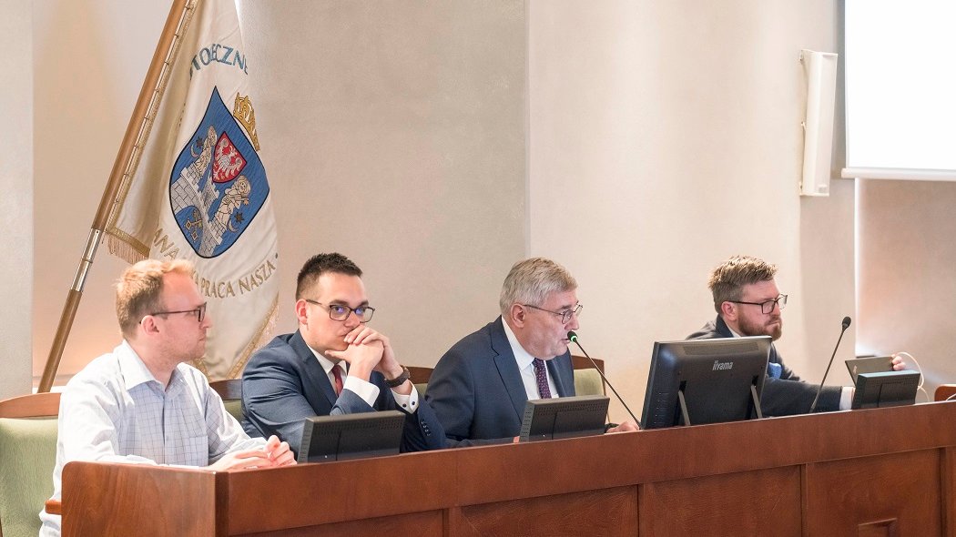 czterech mężczyzn siedzi za stołem prezydilalnym, za nimi sztandar z herbem Poznania