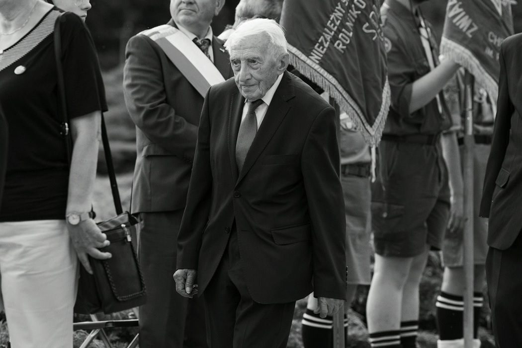 starszy mężczyzna w garniturze, lekko przygarbiony, za nim ludzie stoją w szeregu, niektórzy trzymają sztandary - grafika artykułu