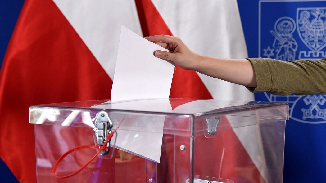 ręka wrzucająca kartkę papieru do przezroczystej urny, za urną flaga polski i herb Poznania - grafika artykułu