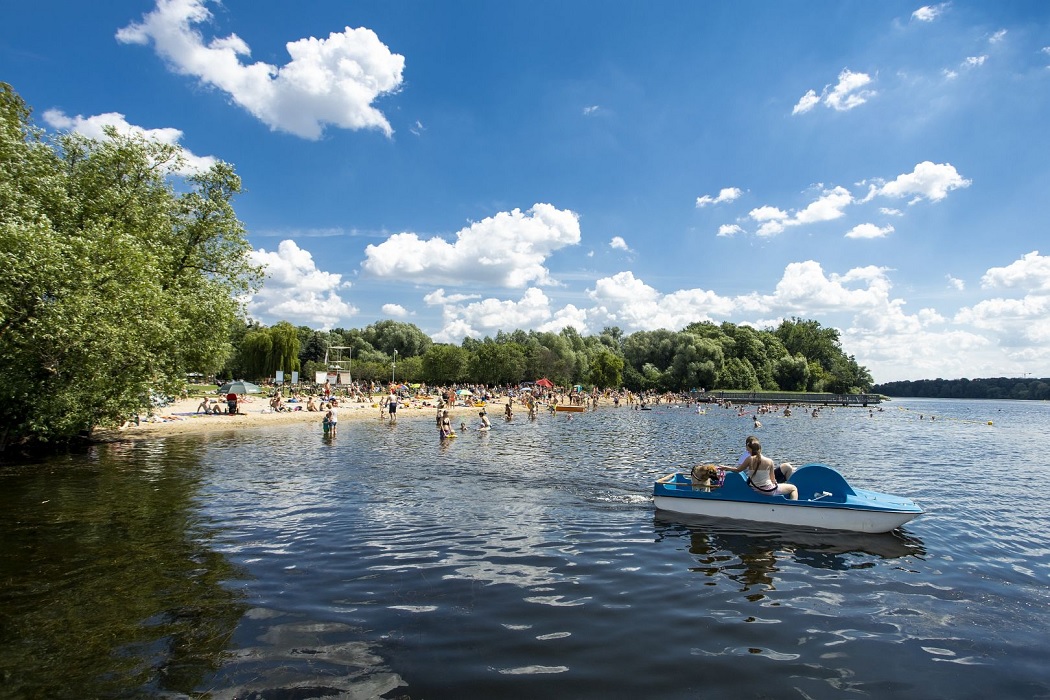 akwen wodny, plaża, ludzie kąpią się lub leżą na plaży, ktoś płynie na rowerze wodnym, plażą otaczają drzewa, niebo jest blękiten, świeci słońsce, trochę jaknych chmurek - grafika artykułu