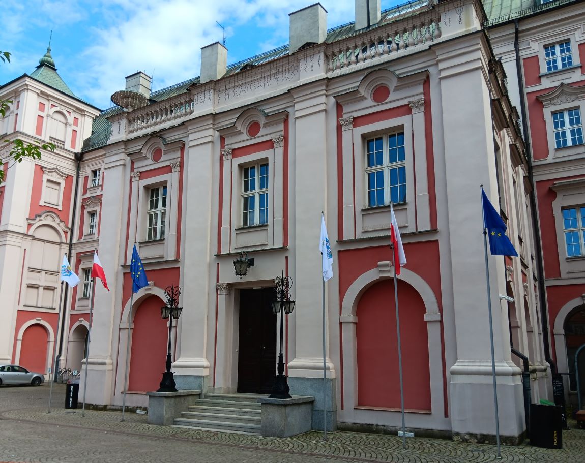 wejście do gmachu Urzędu Miasta Poznania od strony dziedzińca, przed wejściem na masztach z obu stron po trzy flagi: Poznania, Polski i Unii Europejskiej - grafika artykułu