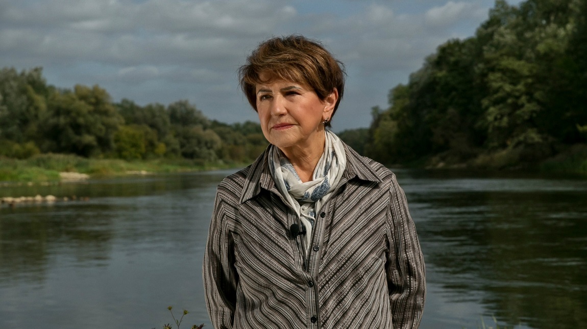 Kobieta ubrana w szarą koszulę i apaszkę, krótkie włosy, stoi na tle jeziora otoczonego gęstą, wysoką roślinnością - grafika artykułu