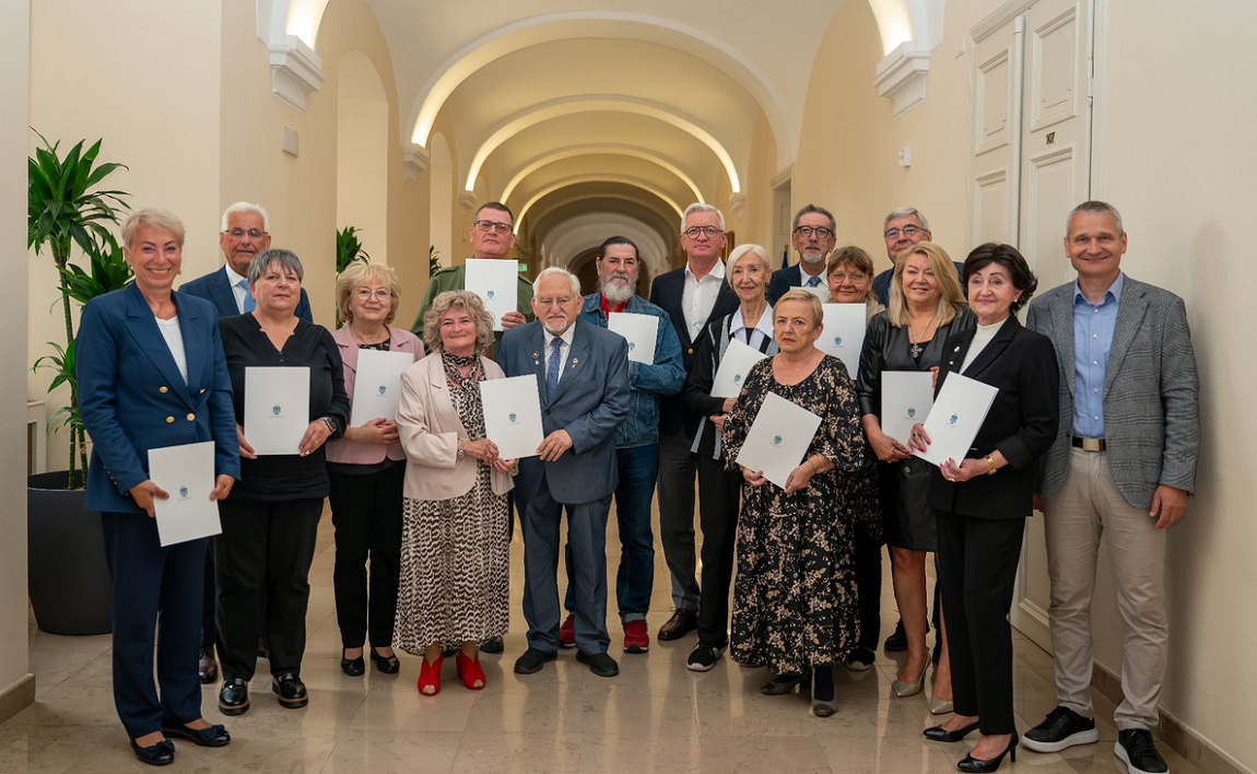 grupa kilkunastu osób, kobie i mężczyzn, większość w wieku senioralnym, pozuje do zdjęcia na korytarzu Urzędu Miasta - grafika artykułu
