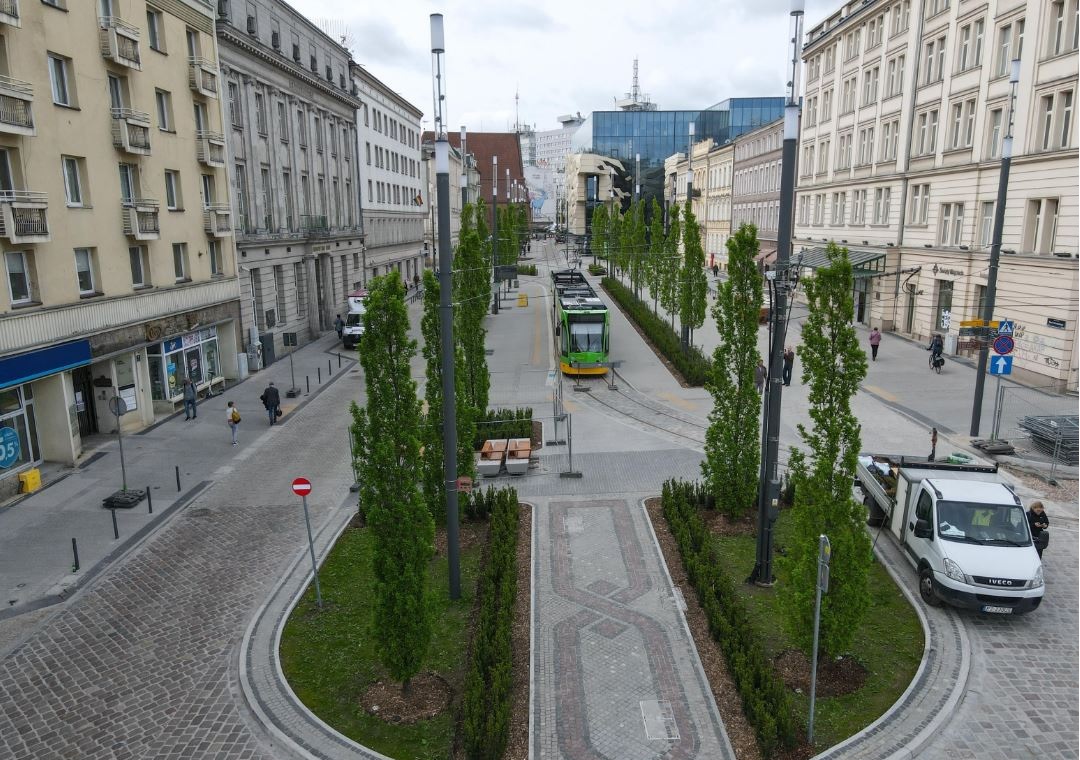 widok na al. Marcinkowskiego z dużej wysokości z okna budynku, widoczne torowisko, drzewa i trawniki dookoła, chodniki, ulice, okoliczne budynki, tramwajo, dwa samochody dostawcze, piesi - grafika artykułu
