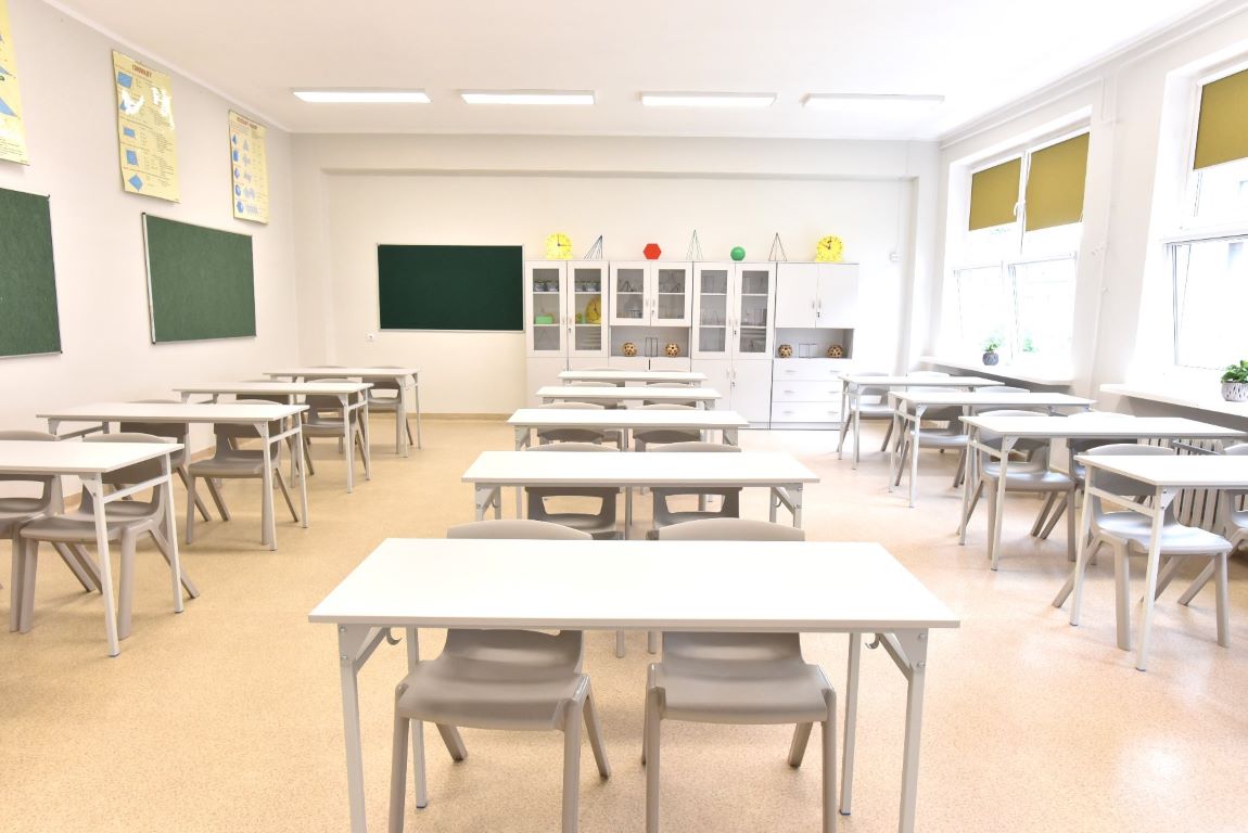 sala lekcyjna, ławki, krzesła i ściany w jasnym kolorze, widać szafki pod ścianą, tablica na ścianie oraz plansze edukacyjne - grafika artykułu