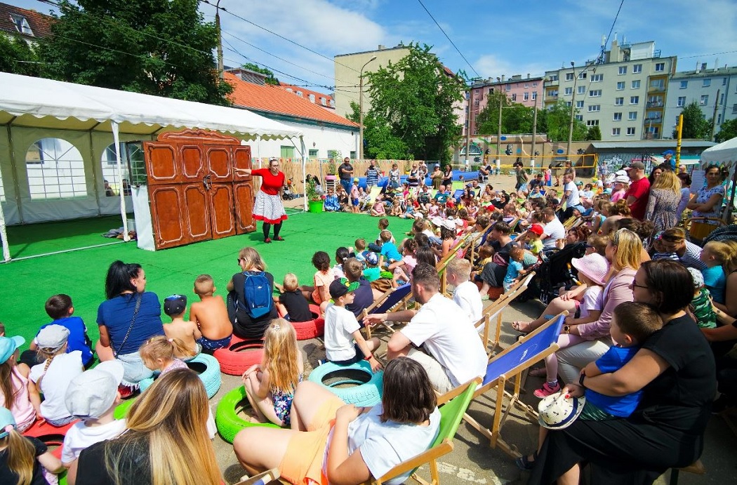 Duża gropa dzieci z rodzicami, są zgomadzeni wokół sceny w plenerze, wśród zabudowy śródmiejskiej, na scenie jedna osoba realizuje spektakl - grafika artykułu