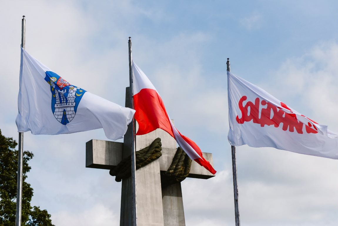 flagi - Poznania, Polski i Solidarności powiewają na masztach na tle nieba, za nimi widoczny fragment pomnika Poznańskiego Czerwca 1956 - grafika artykułu