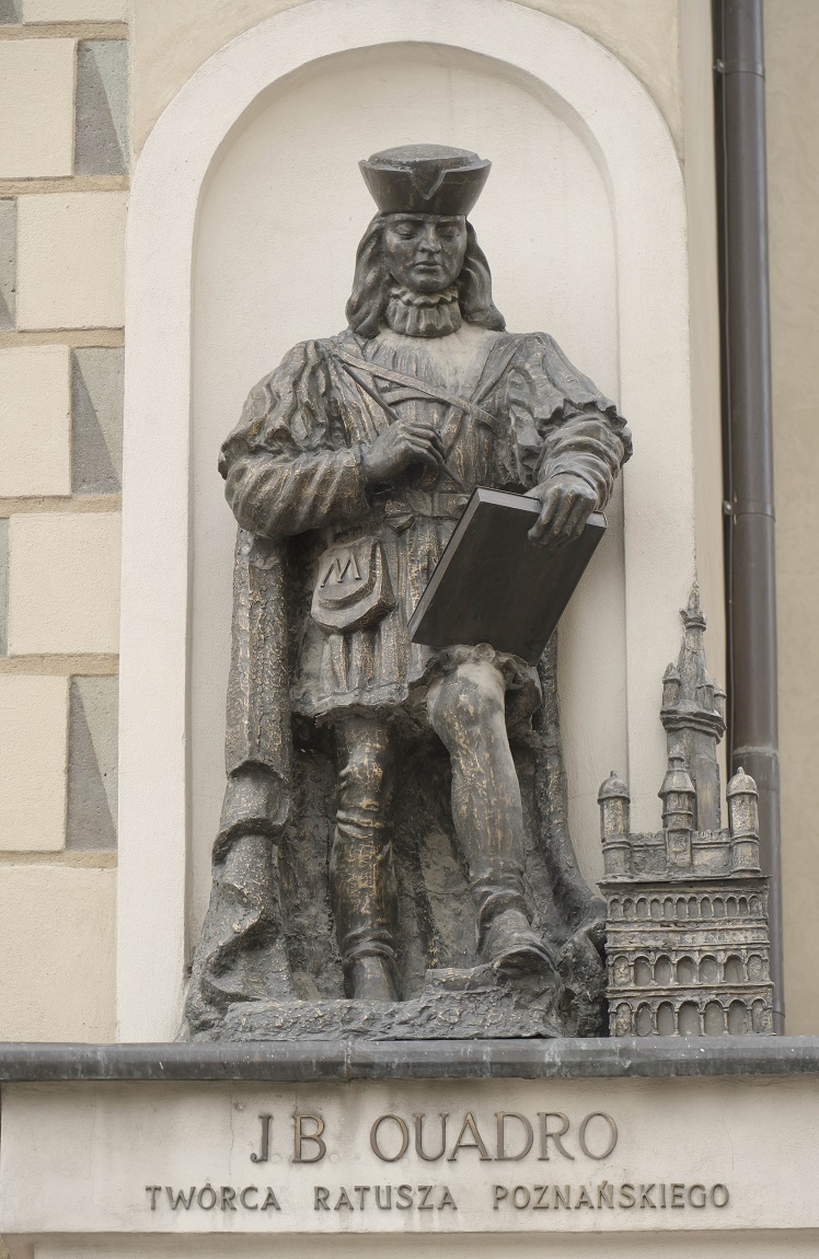 Figura przedstawiająca wizerunek Jana Baptysty Quadro, zdobiąca fasadę kamienicy na narożniku Starego Rynku i ul. Zamkowej, mężczyzna w nakryciu głowy z włosami do ramion ubrany w płaszcz w rękacg trzyma kartkę i coś do pisania, obok niego miniatura ratusza - grafika artykułu