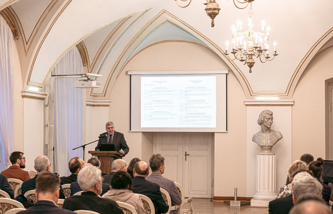 w sali ze zdobionymi łukami siedzą osoby nakrzesłach ustawionych w rzędy, przed nimi jedna osoba stoi przy mównicy, za nią ekran z wyświetloną treścią niewidoczną na zdjęciu - grafika artykułu