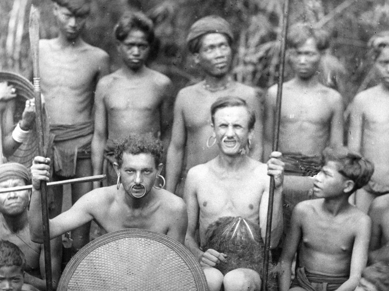 archiwlana fotografia przedstawia mieszkańców obszaru lasów tropikalnych, a między nimi dwóch europejczyków ubranych jak rdzenni mieszkańcy, mają kolczyki w nosie i uszach, trzymają tarcze i włócznie - grafika artykułu