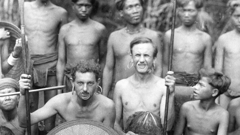 archiwlana fotografia przedstawia mieszkańców obszaru lasów tropikalnych, a między nimi dwóch europejczyków ubranych jak rdzenni mieszkańcy, mają kolczyki w nosie i uszach, trzymają tarcze i włócznie