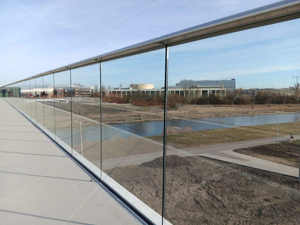 fragment mostu pieszo-rowerowego nad rzeką, most ma balustradę całą z przezroczystego szkła, za nim widoczna rzeka, nadbrzeże i budynku o różnym kształcie i wysokości - grafika artykułu