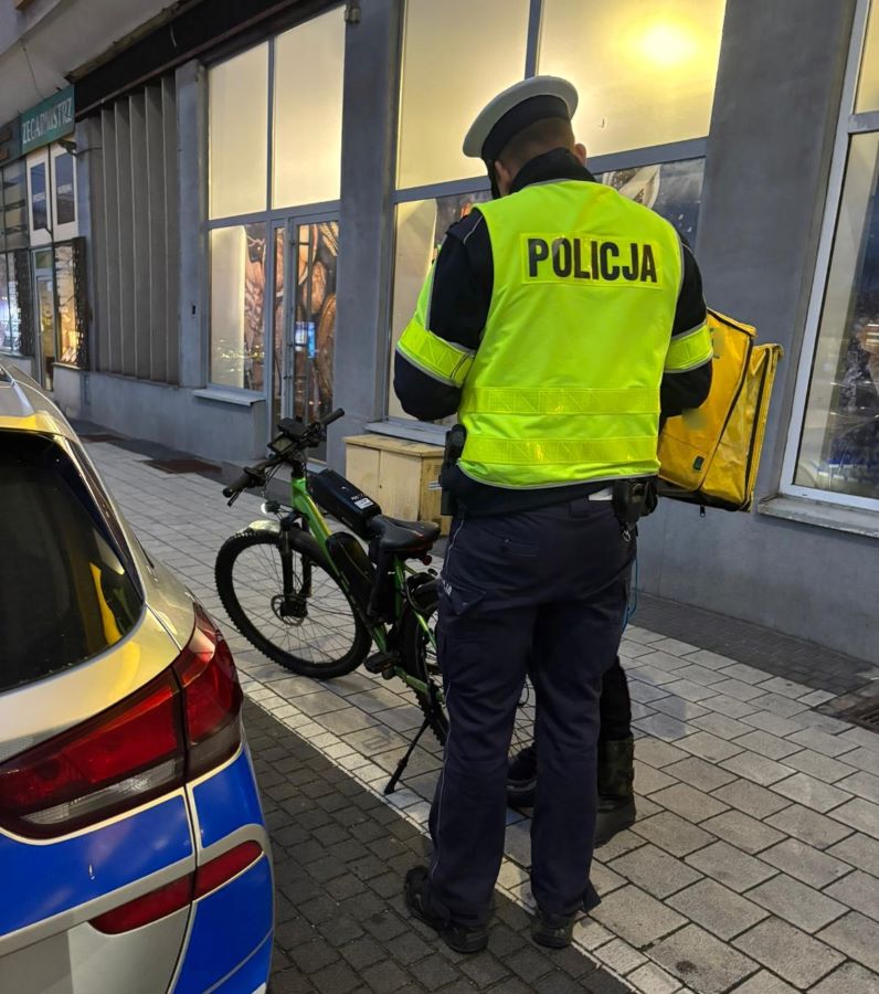 Policjant w odblaskowej kamizelce legitymuje rowerzystę rozwożącego jedzenie, rowerzysta ma charakterystyczny plecak w kształcie kostki, obok widać fragment policyjnego samochodu - grafika artykułu