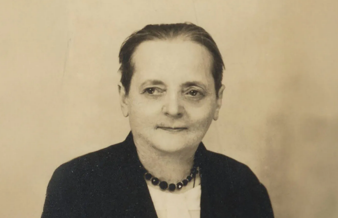 kobieta w średnim wieku, włosy upięte do tyłu, ubrana w czarną suknię, na szyi na naszyjnik z ciemnych kamieni, ma spokojny wyraz twarzy, lekko się uśmiecha - grafika artykułu