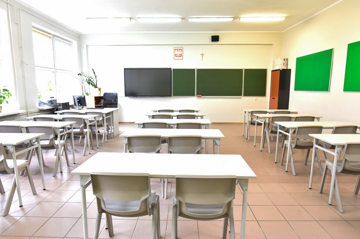 Klasa lekcyjna w szkole, pusta, stoliki i krzesła ustawione w rzędach, po dwa krzesła w ławce, stanowisko dla nauczyciela, na ścianach zielone tablice, w narożniku sali szafa, na ścianie herb Polski i krzyż - grafika artykułu