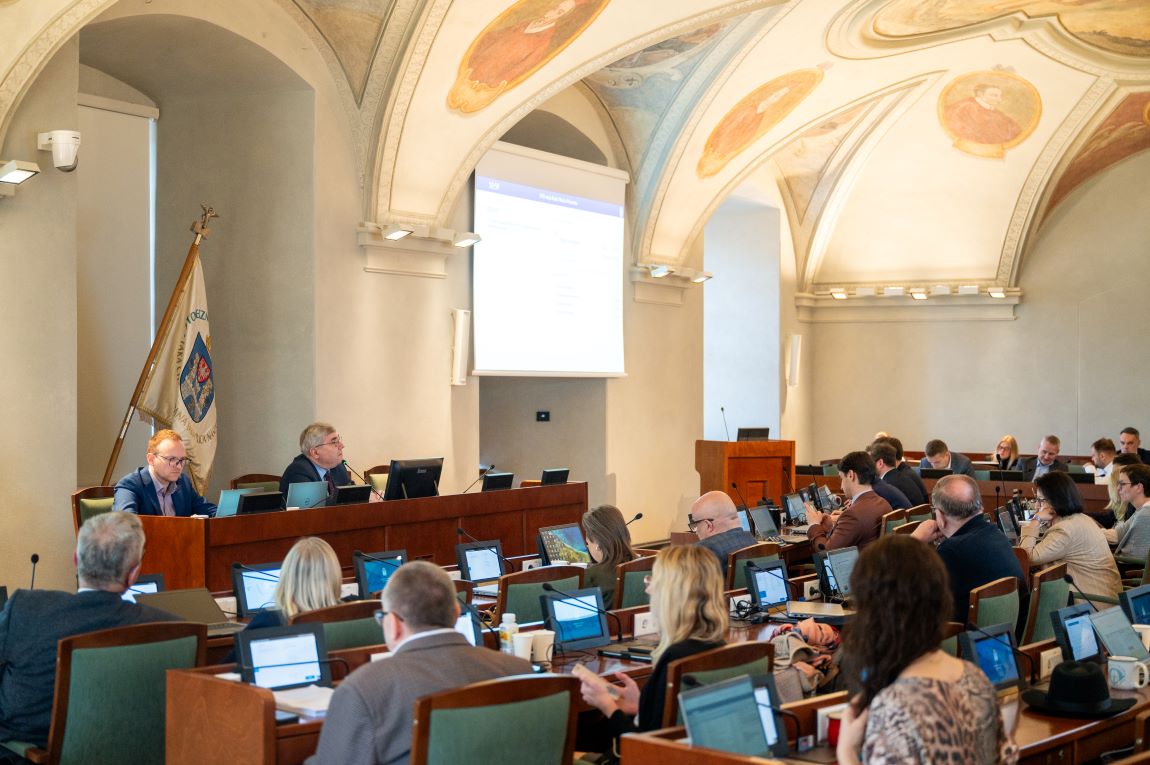 na sali z sufitem zdobionym zabytkowymi freskami obraduje Rada Miasta, za stołem prezydialnym siedzą dwie osoby, przed nimi w rzędach radne i radni siedzą przy swoich stanowiskach, na każdym stanowisku monitor i mikrofon - grafika artykułu