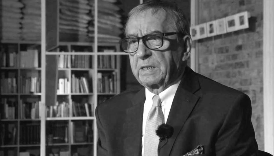 starszy mężczyzna w okularach, krótkie włosy, ujęcie do popiersia, ubrany w ciemną marynarkę, jasną koszulę i krawat, za nim półka z książkami - grafika artykułu