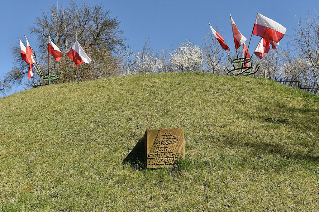 pagórek porośnięty trawą, na nim krzewy, niska roślinnoś, zatknięte biało czerwone flaki, w środku pagórka tablica pamiętkowa - grafika artykułu