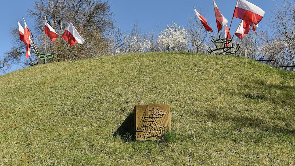 pagórek porośnięty trawą, na nim krzewy, niska roślinnoś, zatknięte biało czerwone flaki, w środku pagórka tablica pamiętkowa