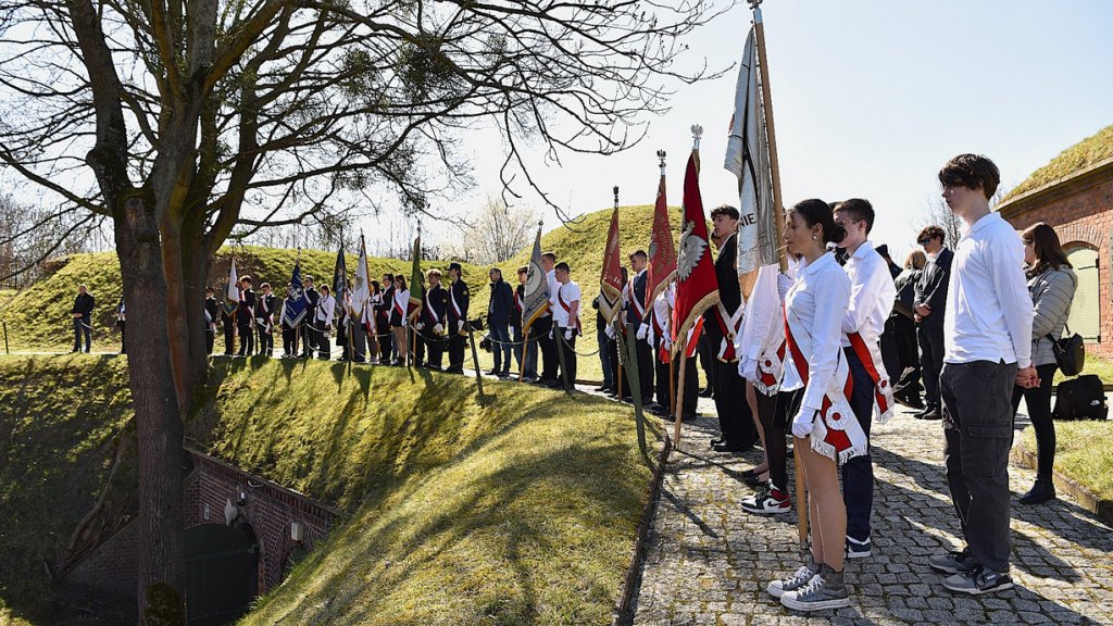 deegacje szkolne z pocztami sztandarowymi, młodzież ubrana na galowo, stoją na ścieżce wokół zabudowań fortecznych