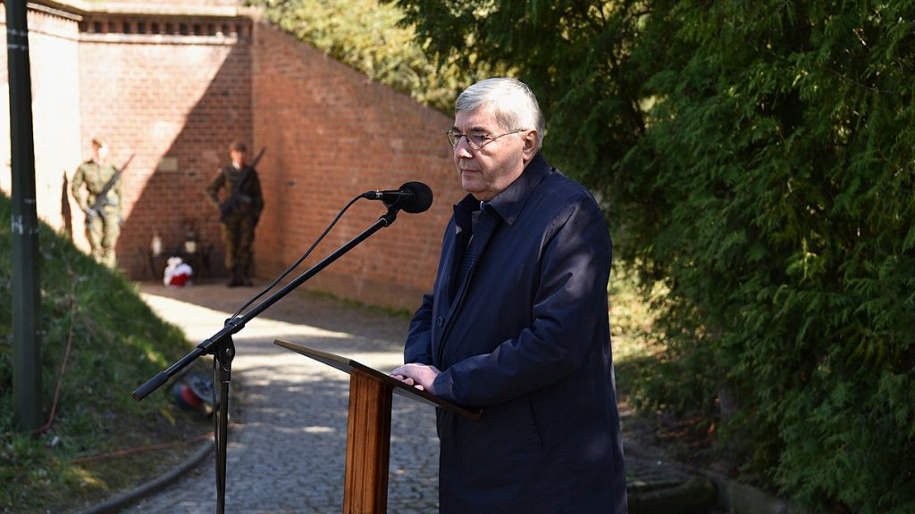 mężczyzna w średnim wieku, siwe włosy, okulary, ubramy w ciemny płaszcz, stoi zamyślony przy mówicu z mikrofonem, za nim zieleń, fragment zabudowy fortyfikacyjnej, w oddali żołnierze pełniący wartę przy tablicy pamiątkowej