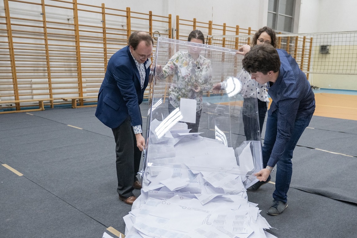 Cztery osoby dwaj mężczyźni i dwie kobiety przeczylają przezroczystą urnę wyborczą wysypując z nieh karty, znajdują się w sali gimnastycznej - grafika artykułu