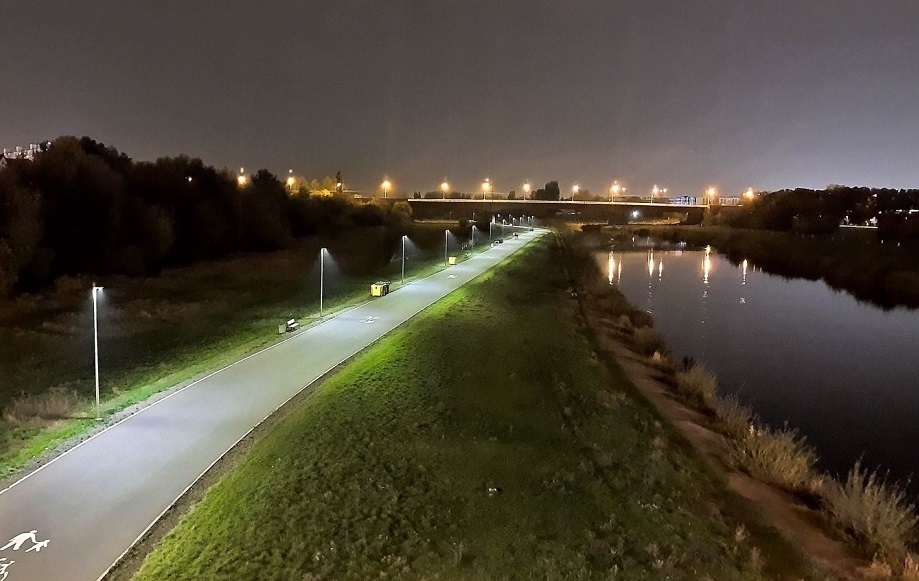 Fragment Wartostrady w porze nocnej, oświetlona ścieżka rowerowa, obok rzeka, na dalszym planie oświetlony most - grafika artykułu