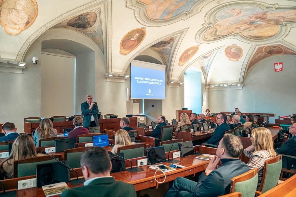 w sali sesyjnej przy stanowiskach z monitoriami i mikrofonami siedzą osoby, przed nimi stoi jedna osoba - mężczyzna trzymający mikrofon, sklepienie sali zdobią kolorowe, zabytkowe freski - grafika artykułu