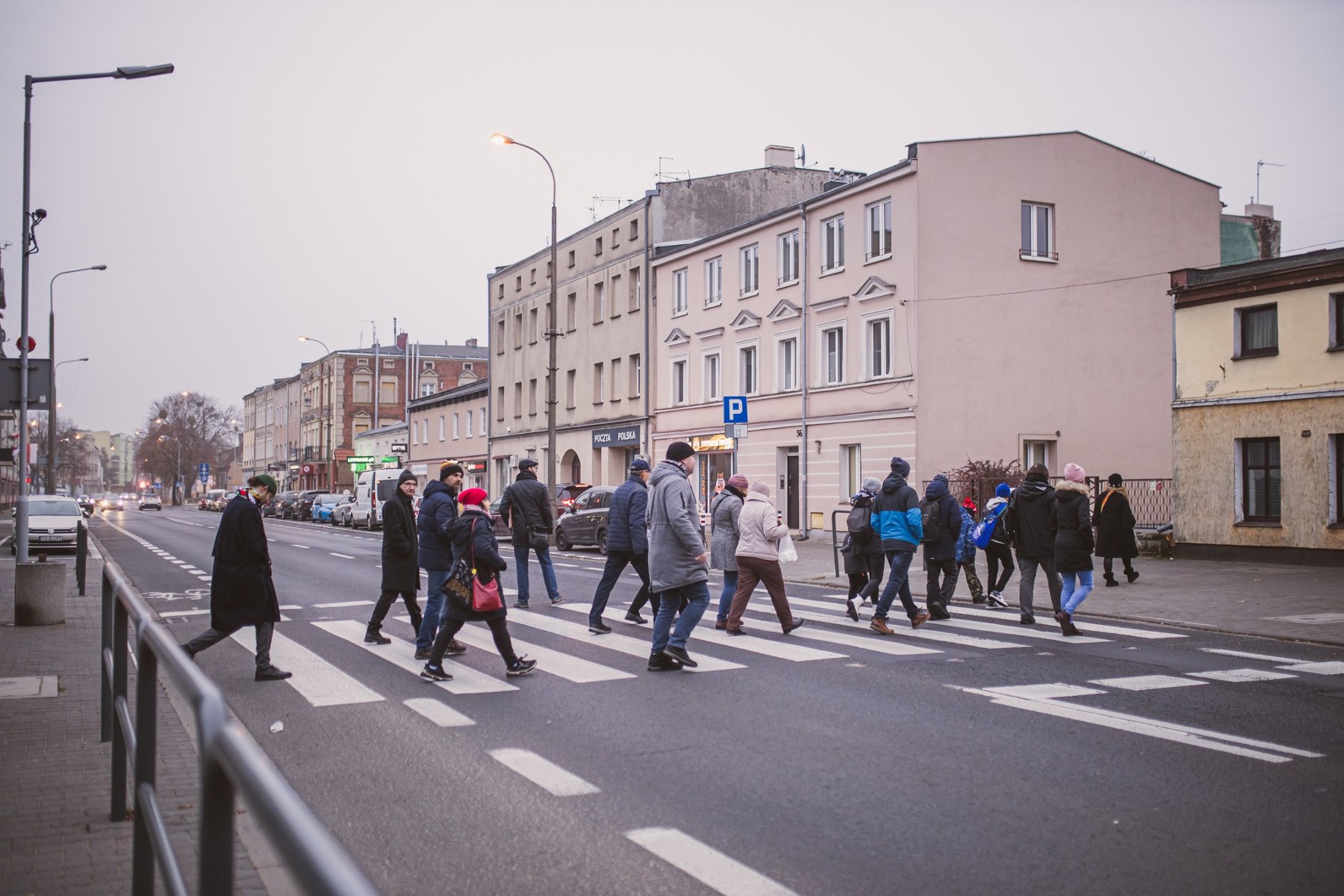 grupa osób przechodzi po pasach przez jezdnię, wzdłuż ulicy stoją dwu i trzy kondygnacyjne kamienice - grafika artykułu