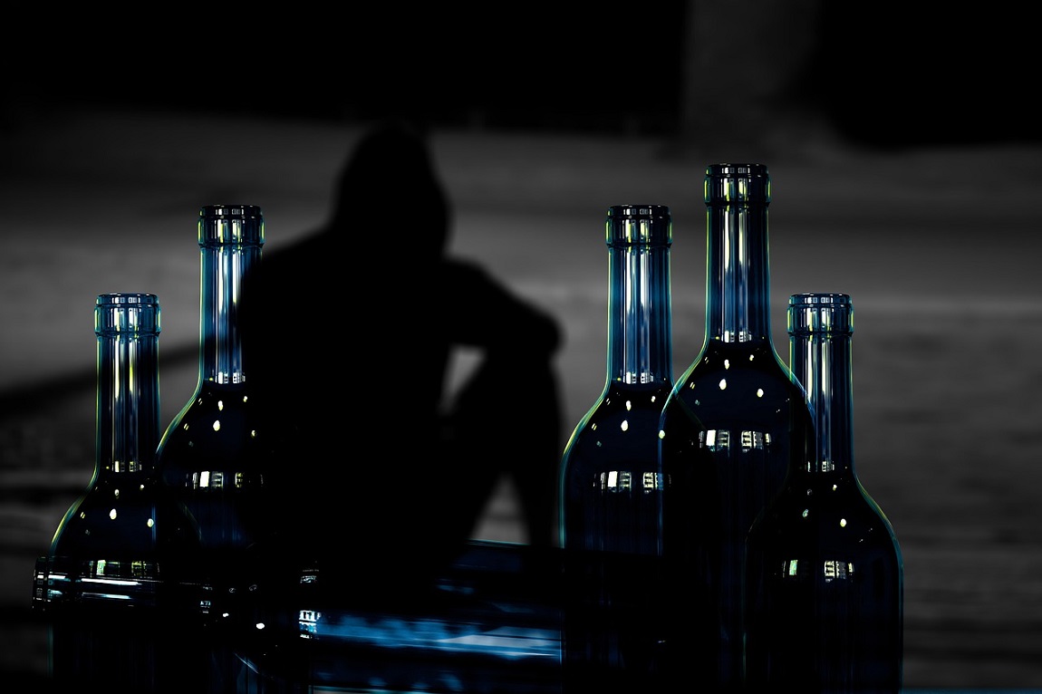 kilka butelek, za nimi ludzka postać w kapturze, odwrócona w pozycji siedzącej, zdjęcie jest zaciemnione - grafika artykułu