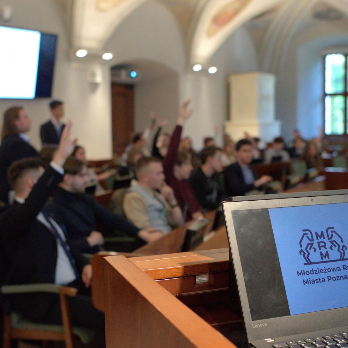 na pierwszym planie otwarty laptop postawiony na mównicy, na ekranie wyświetlone logo i napis Młodzieżowa Rada Miasta Poznania, w tle - zdjęcie zamglone - osoby siedzą przy stanowiska, niektóre z nich mają rękę uniesioną w górę, prawdopodobnie głosują - grafika artykułu