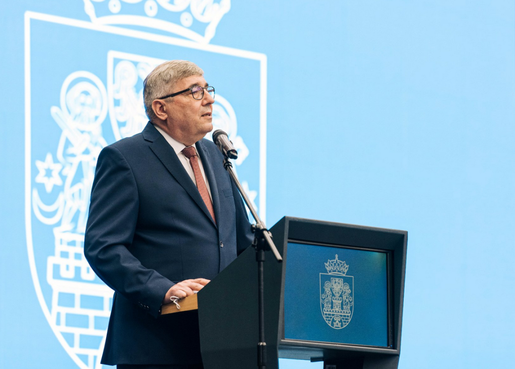 za mówniącą z mikrofonem stoi mężczyzna w garniturze, krawat i jasna koszula, okulary, włosy krótkie zaczesane na bok, częściowo siwiejące, z przodu mównicy herb Miasta Poznania, za mężczyzną ściana z dużym herbem Poznania - grafika artykułu