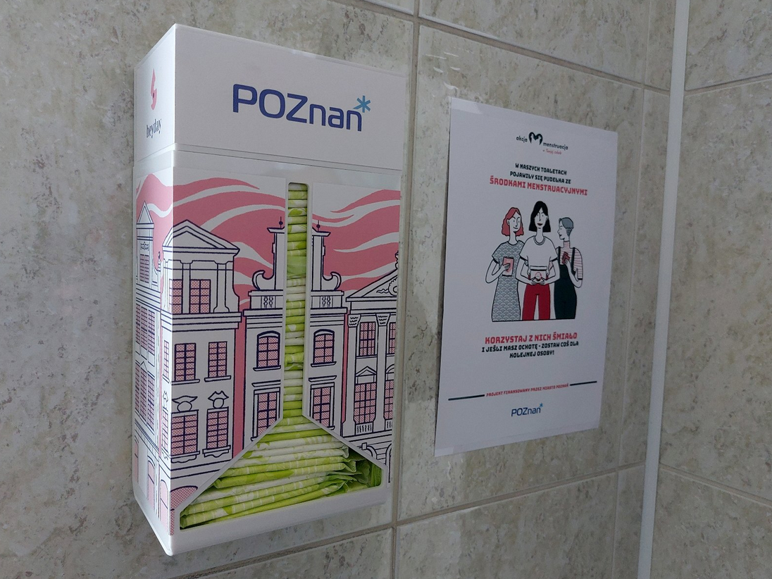 na ścianie wyłożonej kafelkami zamontowana biało różowa skrzynka z logo Poznania i grafiką, w niej środki higieniczne, obok przyklejony do ściany edukujący o środkach higieny menstruacyjnej - grafika artykułu