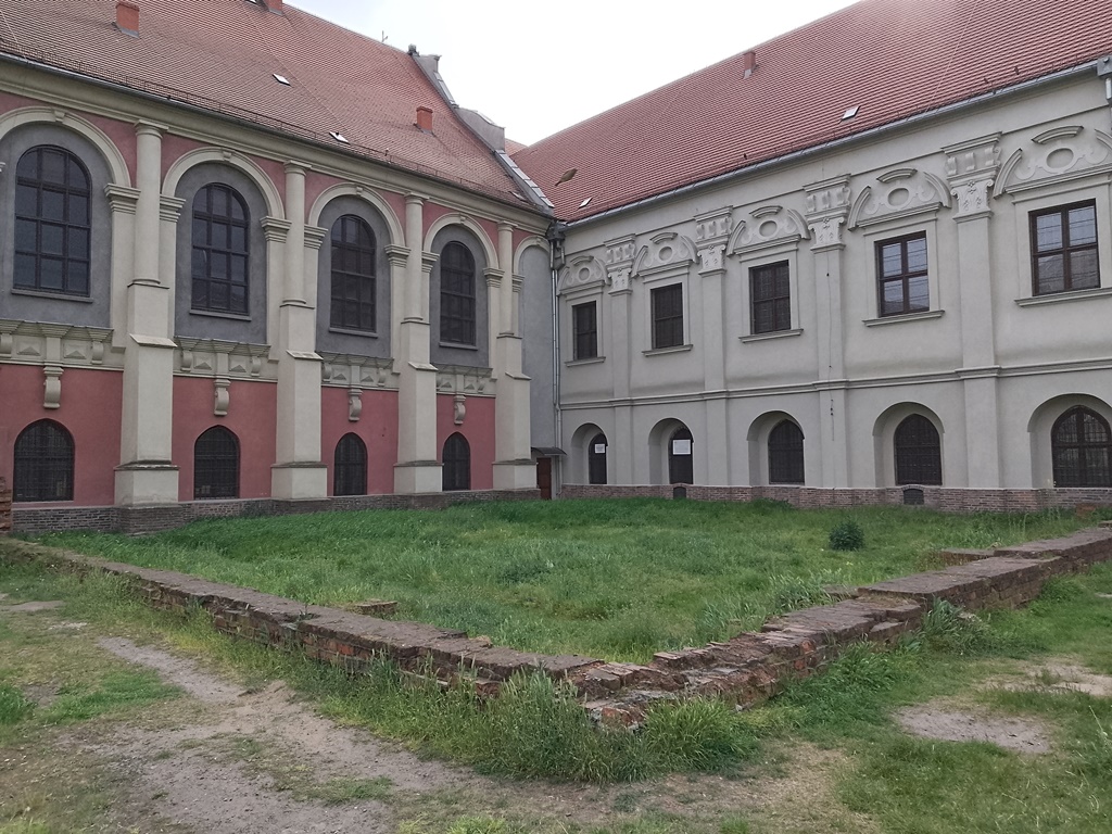 dwa zabytkowe budynki ze spadzistymi dachami, wysokimi oknami zdobionymi łukami, przed nimi teren porośnięty trawą i pozostałości murów lub ścian budynków - grafika artykułu