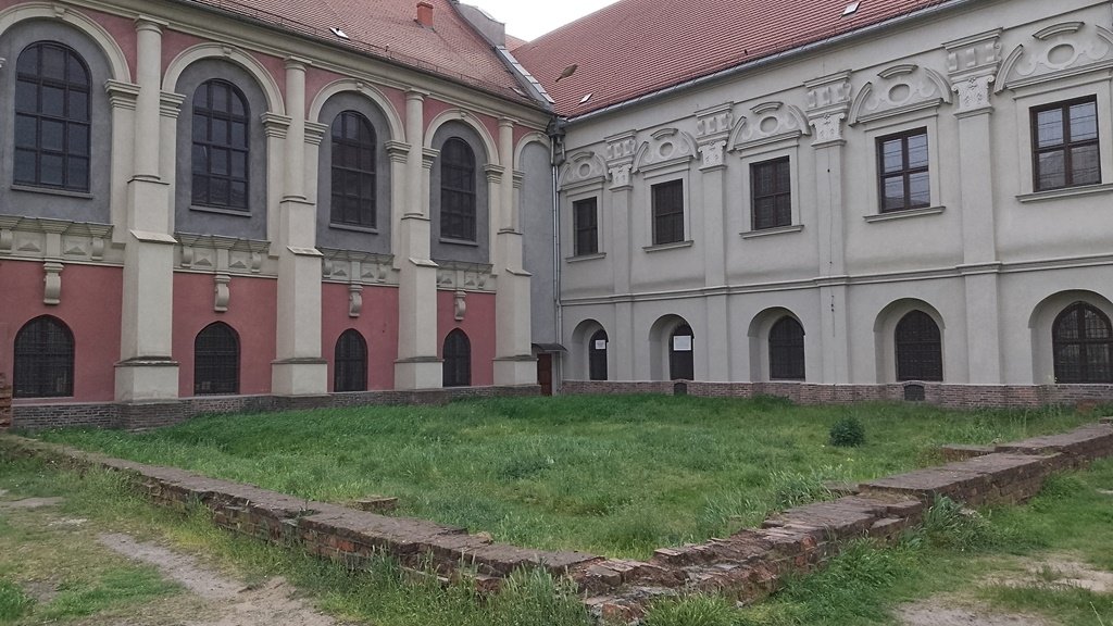 dwa zabytkowe budynki ze spadzistymi dachami, wysokimi oknami zdobionymi łukami, przed nimi teren porośnięty trawą i pozostałości murów lub ścian budynków