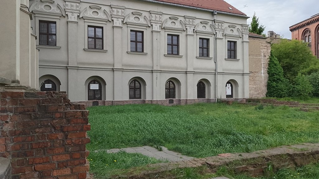 zabytkowy budynek ze spadzistym dachem, wysokimi oknami zdobionymi łukami, przed nim teren porośnięty trawą i pozostałości murów lub ścian budynków