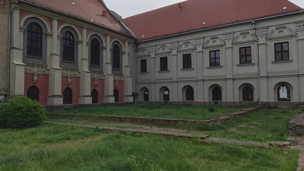 dwa zabytkowe budynki ze spadzistymi dachami, wysokimi oknami zdobionymi łukami, przed nimi teren porośnięty trawą i pozostałości murów lub ścian budynków