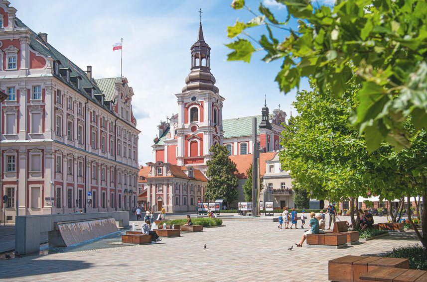 wybrukowany skwer, ludzie siedzą na ławkach lub spacerują, w tle wzniosłe gmachy, na jednym z nich powiewa biało-czerwona flaga - grafika artykułu