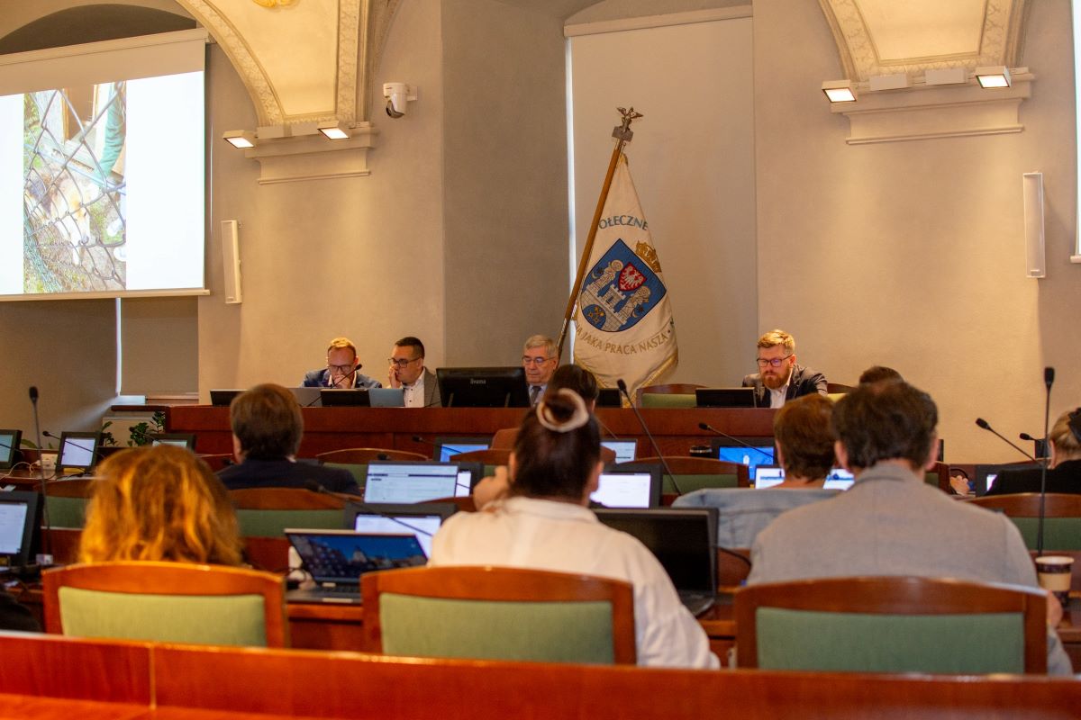 w sali zdobionej freskami widać stół prezydialny przy którym siedzą cztery osoby, przed nimi rzędy siedzeń, przy których też siedzą osoby - grafika artykułu