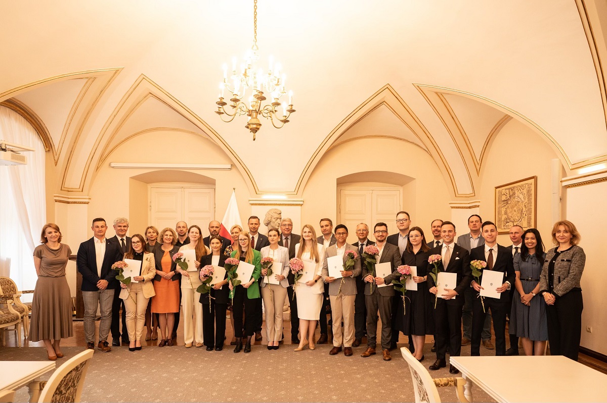 grupa osób kobiet i mężczyzn w różnym wieku pozuje do wspólnego zdjęcia, są elegancko ubrani, niektórzy z nich trzymają kwiaty, znajdują się w eleganckiej, zabytkowej sali z żyrandolem - grafika artykułu